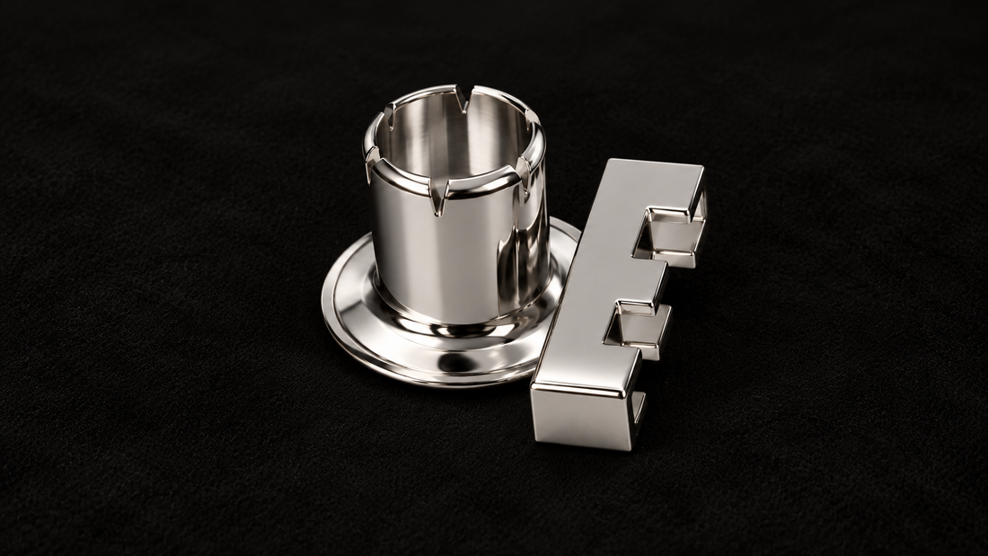 KORTONS platinum Letter E Signature Eyelet displayed alongside its cylindrical mounting barrel, featuring a polished precious-metal finish on a smooth, all-black suede background.