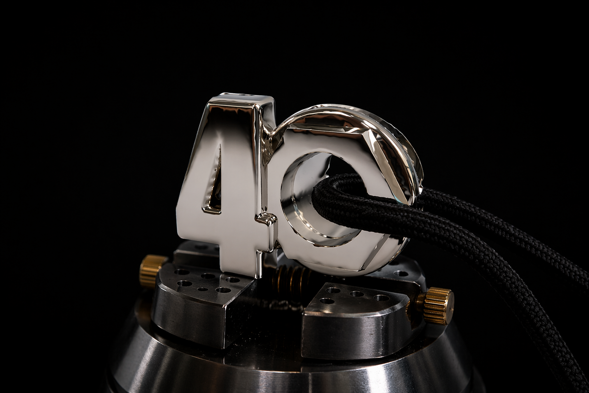 Black shoelace with aglet threaded through an octagonal metal eyelet in a polished silver “40” pendant, secured on a precision jewelry vise against a dark luxury background.