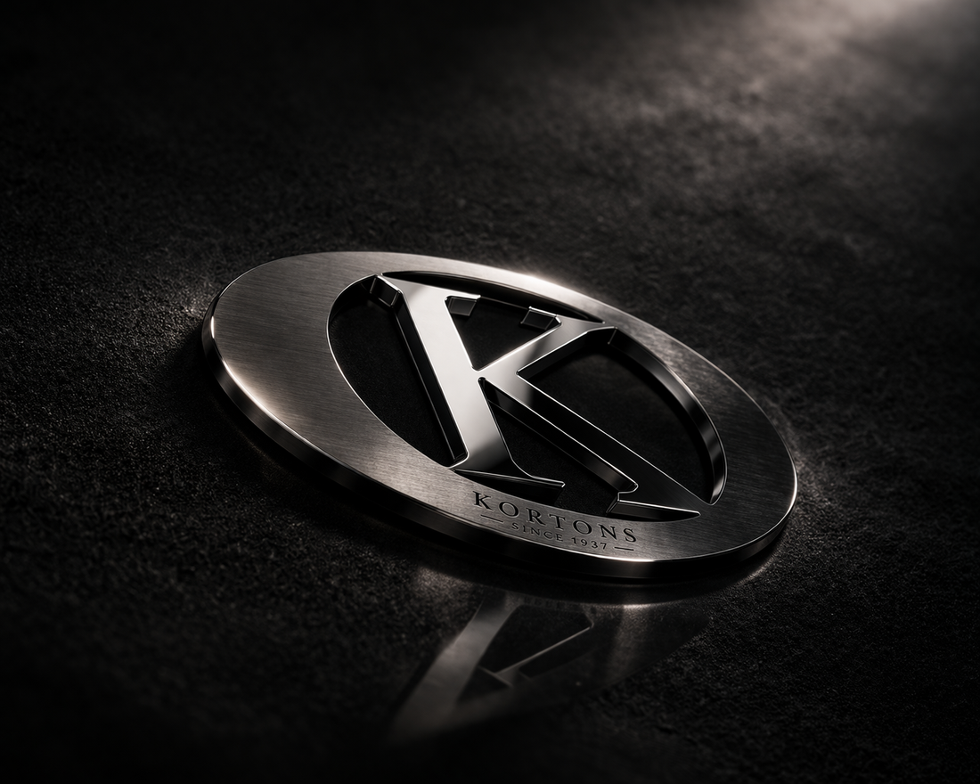 Blackened metal KORTONS emblem with engraved “Since 1937” resting on a dark suede-textured surface with subtle reflection and dramatic lighting, showcasing a refined oval badge and polished “K” centerpiece