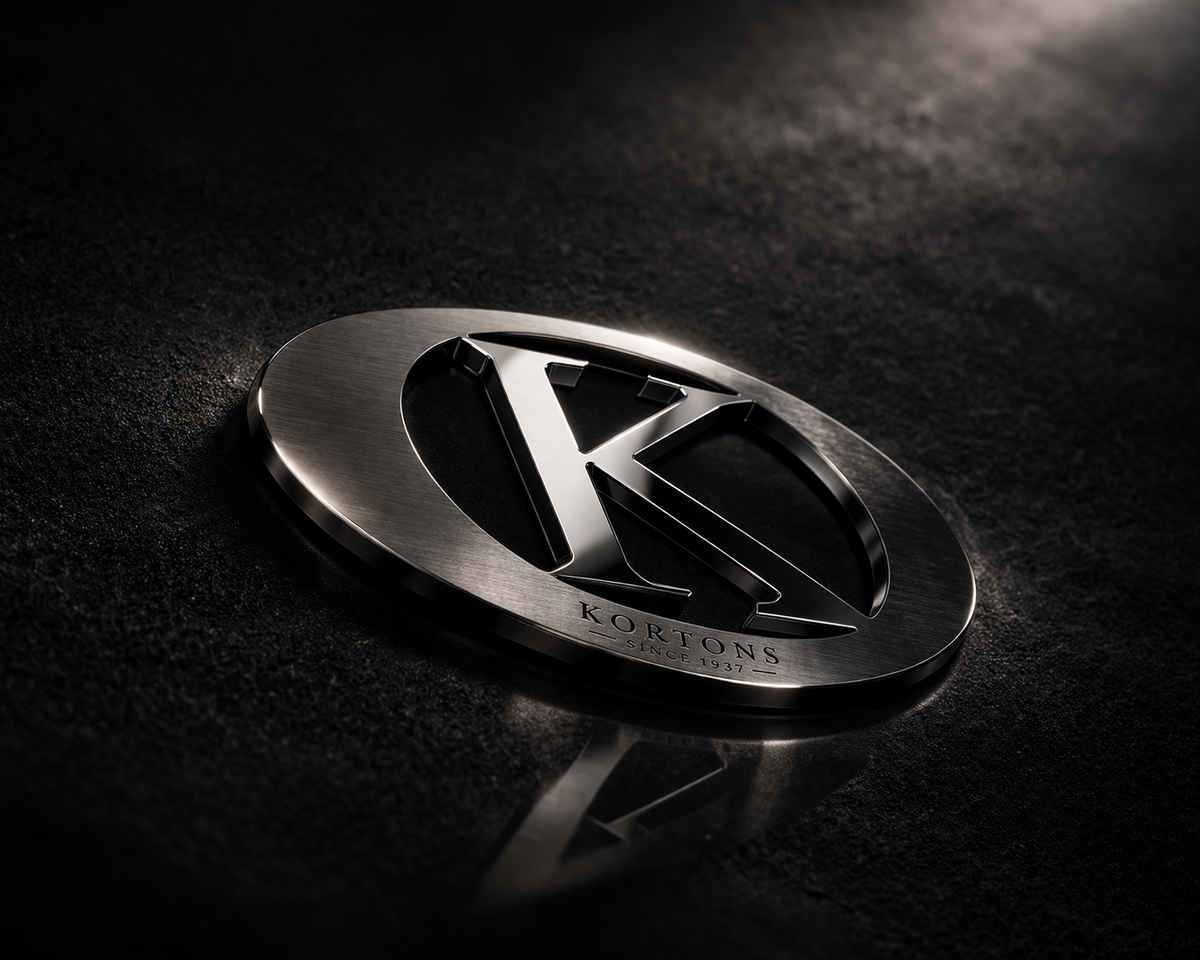 Blackened metal KORTONS emblem with engraved “Since 1937” resting on a dark suede-textured surface with subtle reflection and dramatic lighting, showcasing a refined oval badge and polished “K” centerpiece