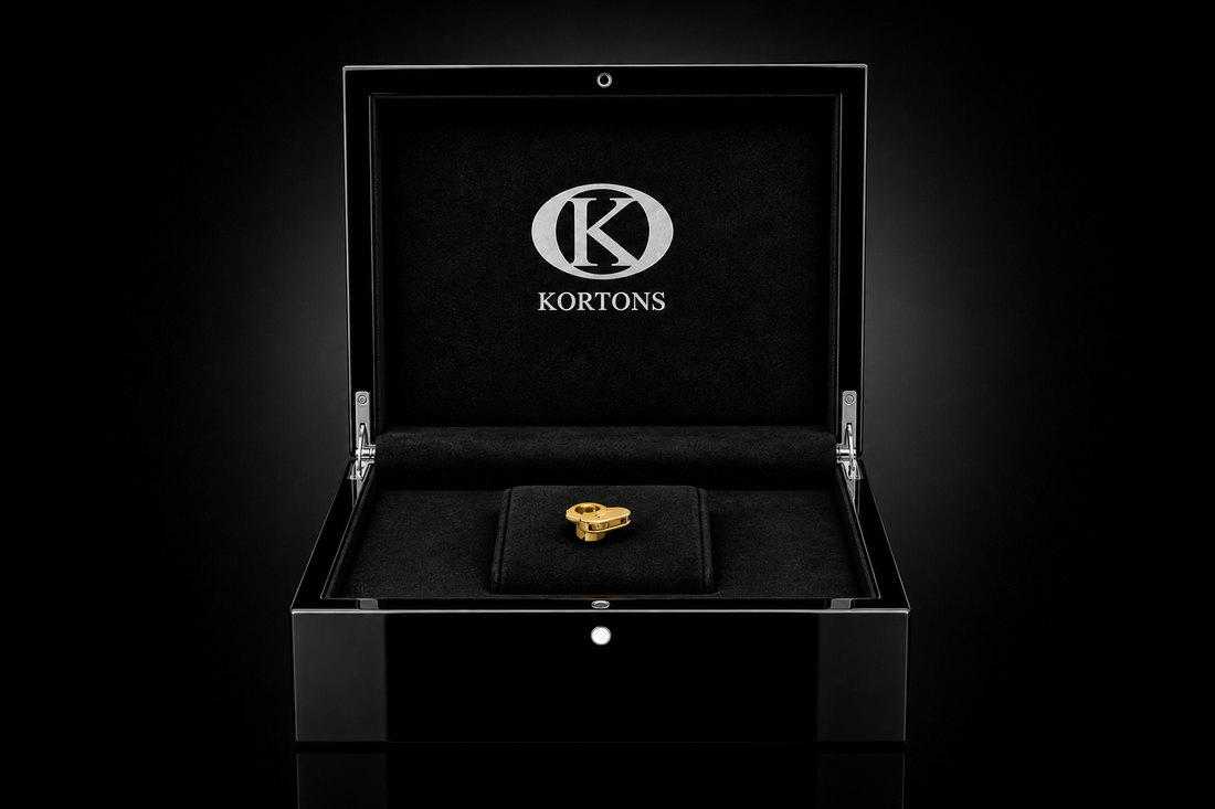 Front-facing KORTONS glossy black presentation box with black suede interior, displaying a single gold eyelet centered on a raised cushion, set against a subtle black gradient reflective background.