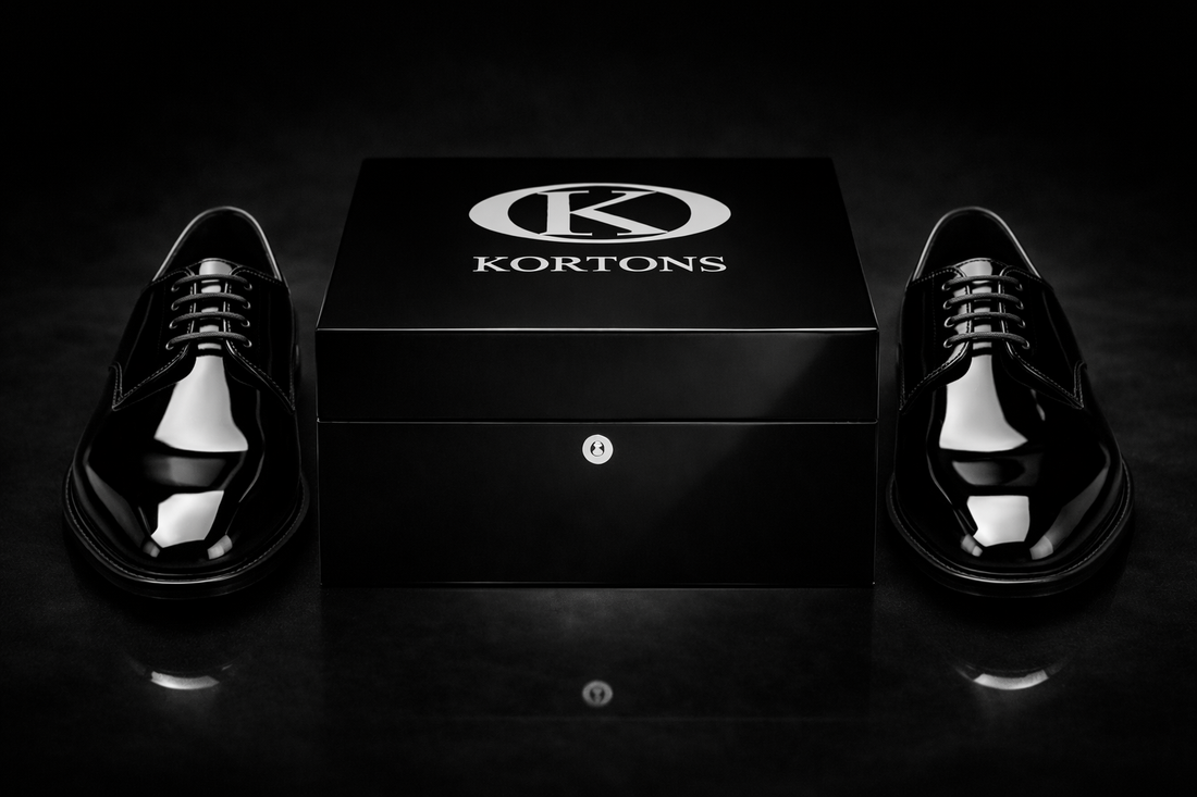 Pair of black patent leather derby tuxedo shoes with high-gloss finish positioned symmetrically beside a Kortons luxury shoe box on a black suede background, highlighting refined lacing, aglets, and eyelets with cinematic lighting and subtle reflections