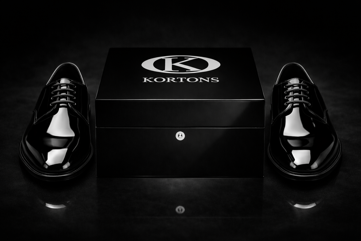 Pair of black patent leather derby tuxedo shoes with high-gloss finish positioned symmetrically beside a Kortons luxury shoe box on a black suede background, highlighting refined lacing, aglets, and eyelets with cinematic lighting and subtle reflections