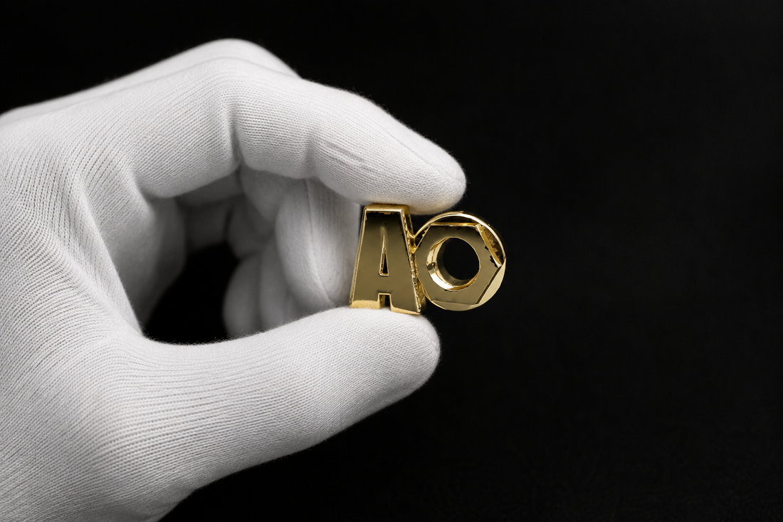 White-gloved hand delicately holding a polished gold KORTONS alphabetical shoe eyelet against a deep black background with dramatic luxury studio lighting and reflective metallic detailing.