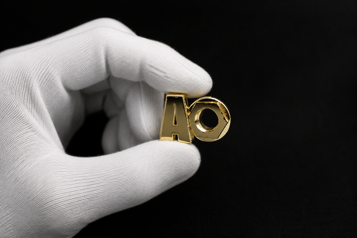 White-gloved hand delicately holding a polished gold KORTONS alphabetical shoe eyelet against a deep black background with dramatic luxury studio lighting and reflective metallic detailing.
