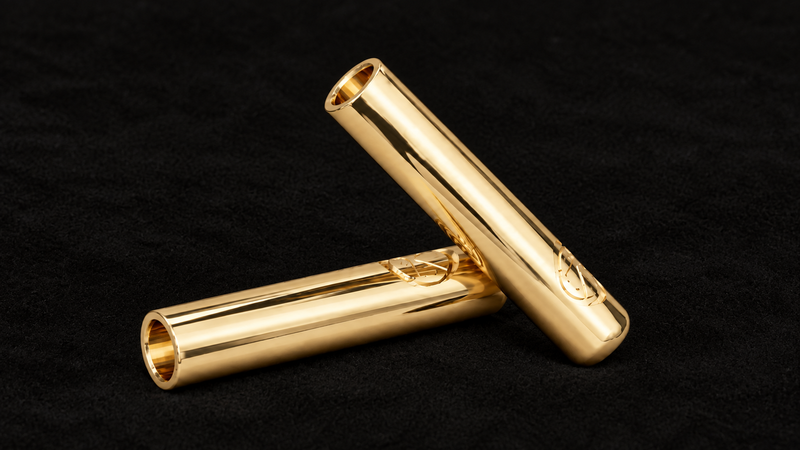 Two polished 14 karat gold Kortons aglets arranged together on a smooth all-black suede background, showcasing their reflective finish and engraved detailing.
