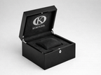 Open KORTONS black luxury eyelet box with glossy exterior and black suede interior, featuring a centered cushion insert and engraved K logo, presented in a clean, minimal studio setting.